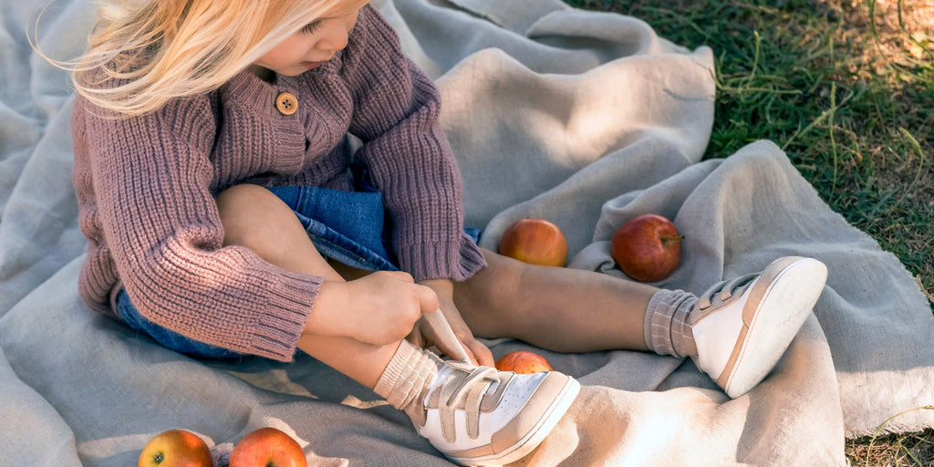 niña atándose los zapatos muris (modelo petra) sentada encima de una manta y con manzanas alrededor
